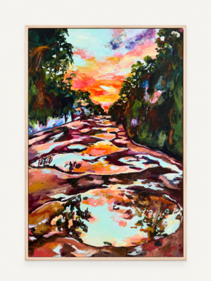 Een schilderij in een lichte houten lijst met een warme, kleurrijke compositie die een pad of weerspiegeling in water bij zonsondergang of zonsopgang toont, geflankeerd door bomen.